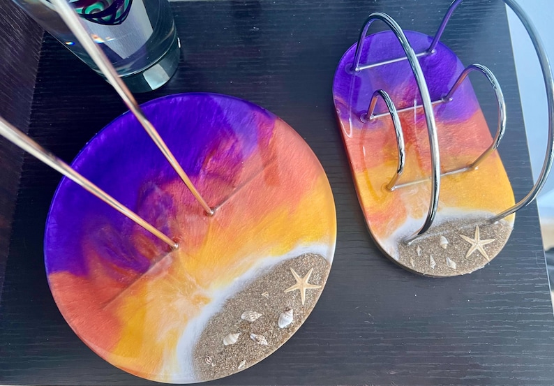May include: Two round resin coasters with a sunset design. The coasters are purple, orange, and yellow with white sand and seashells. One coaster has a metal holder for incense sticks.