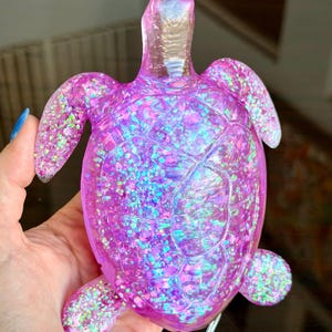 May include: A translucent pink resin turtle figurine featuring a shell filled with iridescent, multi-colored glitter. The turtle has a smooth, glossy finish with visible molded shell segments and a clear, reflective head and flippers.