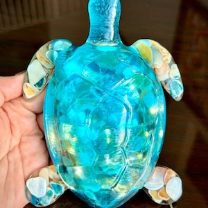 May include: A translucent sea turtle figurine crafted from bright blue resin. The shell and flippers are embedded with various small, smooth, multi-colored seashells in shades of white, tan, and light blue, creating a textured, mosaic-like appearance.