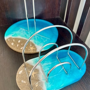 May include: A set of two blue and brown resin napkin holders with a beach theme. The holders are made of resin and have a silver metal frame. The holders are shaped like a beach scene with waves, sand, and seashells.
