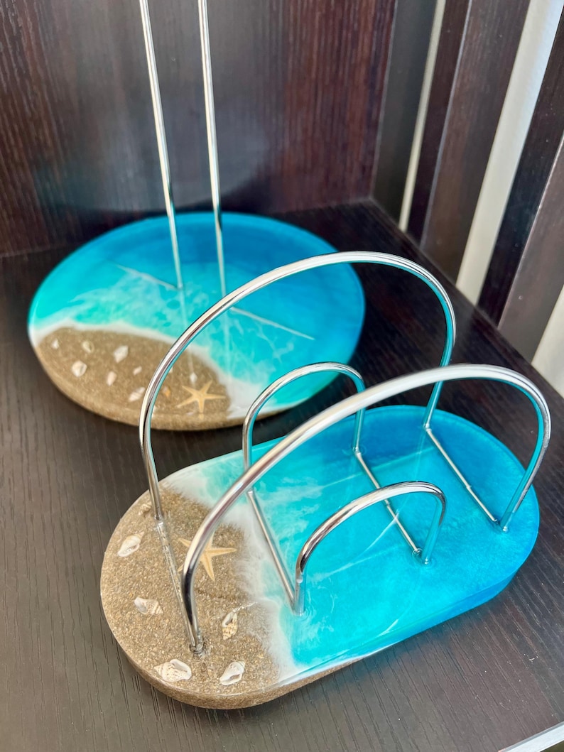 May include: A set of two blue and brown resin napkin holders with a beach theme. The holders are made of resin and have a silver metal frame. The holders are shaped like a beach scene with waves, sand, and seashells.