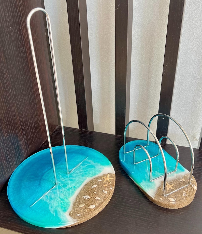 May include: Two resin napkin holders with a beach theme. One holder is a round dish with a blue and white wave design and a starfish. The other holder is a silver wire napkin holder with a blue and white wave design and a starfish.