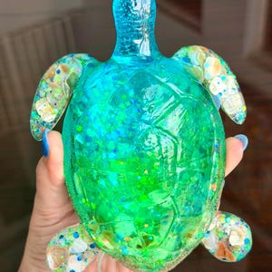 May include: A translucent sea turtle figurine crafted from resin in vibrant shades of blue and green. The shell and flippers are embedded with iridescent glitter and small, light-colored shell fragments, creating a shimmering, multi-textured appearance.