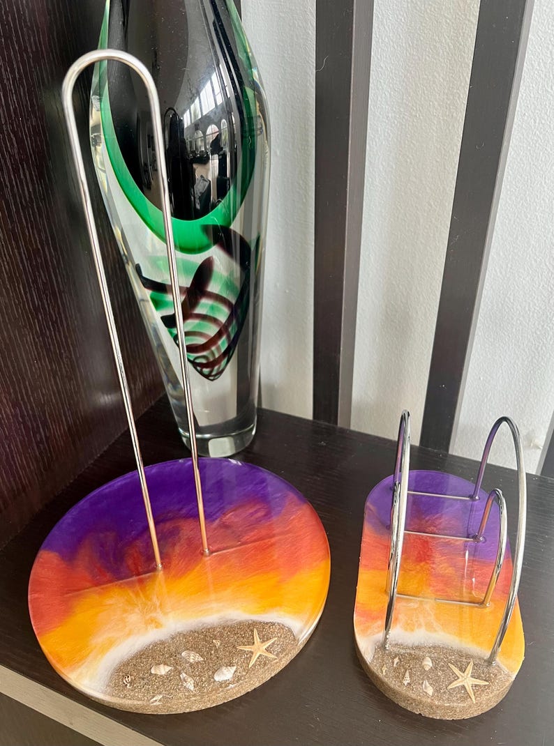 May include: Two resin paper towel holders with a sunset design. The holders are round and have a white sandy beach base with seashells and a starfish. One holder is for paper towels and the other is for napkins.