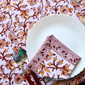 Może przedstawiać: Biały ceramiczny talerz obiadowy z złożonym serwetką na wierzchu. Serwetka jest wykonana z białej tkaniny z czerwono-żółtym wzorem kwiatowym. Talerz znajduje się na białym obrusie z czerwono-żółtym wzorem kwiatowym.