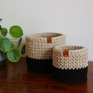 Może przedstawiać: Dwukolorowe kosze z szydełka w kolorze beżowym i czarnym, z małą skórzaną metką. Kosze są różnej wielkości i umieszczone na drewnianej powierzchni. Po lewej stronie znajduje się roślina doniczkowa z okrągłymi zielonymi liśćmi.