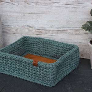 Peut inclure: Un panier de rangement en crochet bleu sarcelle avec une base en cuir marron. Le panier est posé sur une surface noire.