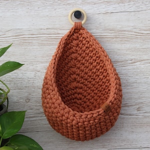 Peut inclure: Un pot de fleurs suspendu en crochet de couleur terre cuite avec un anneau en bois pour l'accrocher. Le pot est vide et prêt à accueillir une plante.