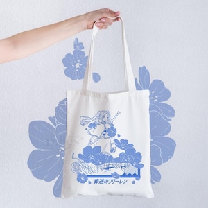 May include: Cream-coloured tote bag featuring a blue anime-style illustration of a character with a sword and floral elements. Japanese text is printed at the bottom. The bag is held by a hand against a white wall decorated with blue flower patterns.