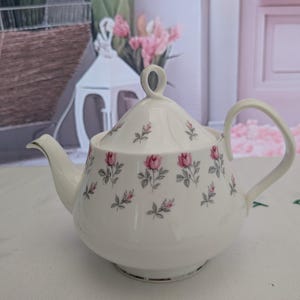 May include: A cream-coloured porcelain teapot featuring a delicate pattern of small pink rosebuds with grey leaves. The teapot includes a matching lid with a loop-shaped handle, a curved spout, and a rounded handle, all accented with a thin silver trim.