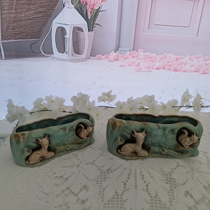 May include: Two oval-shaped ceramic planters featuring a textured, light green finish that mimics tree bark. Each planter is adorned with a small, sculpted beige deer resting on one side and a squirrel perched on the other side of the exterior.