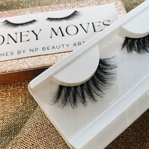 Könnte beinhalten: Zwei Paar falsche Wimpern in einem weißen Plastik-Etui. Auf dem Etui steht "MONEY MOVES" und "LASHES BY NP BEAUTY ART".