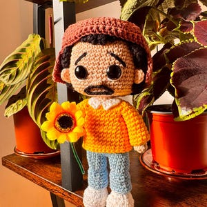 Pode incluir: Uma boneca de crochê feita à mão representando uma pessoa com cabelo escuro, bigode e um gorro cor de ferrugem. A boneca veste um suéter amarelo brilhante de manga comprida com gola branca e calças azul-claras, segurando um girassol.