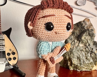 May include: A crochet doll with brown hair, large black eyes, and a light blue shirt. The doll is holding a small tan guitar with white strings and is wearing tan pants with black and white shoes. The figure has a large head and small body.