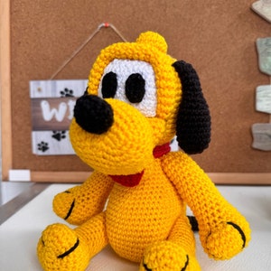 May include: A crocheted plush toy dog in yellow with black ears, nose, and eyes. The dog is wearing a red collar and is sitting on a white surface.