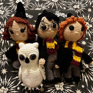 May include: Four crocheted stuffed toys, including a white owl, a brown-haired girl wearing a witch's hat, a boy with black hair wearing glasses and a red and gold scarf, and a boy with brown hair wearing a black robe and a yellow scarf.