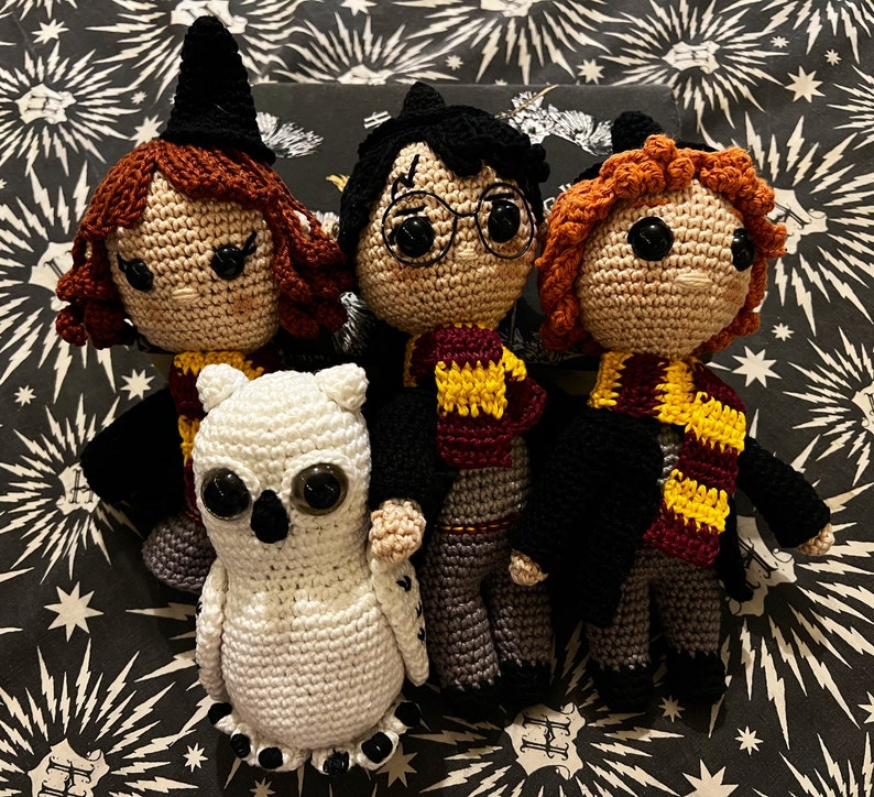 May include: Four crocheted stuffed toys, including a white owl, a brown-haired girl wearing a witch's hat, a boy with black hair wearing glasses and a red and gold scarf, and a boy with brown hair wearing a black robe and a yellow scarf.