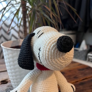 以下が含まれることがあります： 黒色の耳と鼻を持つ、白の毛糸で編まれたぬいぐるみ犬。犬は後ろに傾いた頭で座っており、赤い首輪をしています。