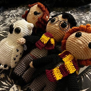 May include: Crocheted Harry Potter characters, including Harry, Ron, Hermione, and Hedwig the owl. The characters are wearing their Hogwarts house scarves and are sitting on a black and white patterned fabric.