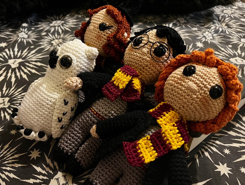 May include: Crocheted Harry Potter characters, including Harry, Ron, Hermione, and Hedwig the owl. The characters are wearing their Hogwarts house scarves and are sitting on a black and white patterned fabric.