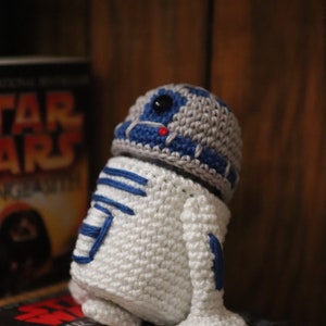 Könnte beinhalten: Ein gehäkeltes R2-D2-Spielzeug mit weißem Körper, blauen und grauen Details und einem schwarzen Auge. Das Spielzeug sitzt auf einem Buch mit dem Titel "Heir to the Empire".