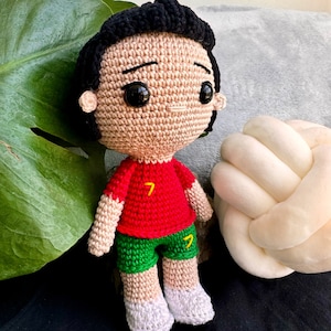 Puede incluir: Muñeco amigurumi de crochet con un jersey de fútbol rojo y verde con el número 7 en la parte delantera. El muñeco tiene el pelo negro y una expresión neutral.