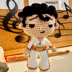 May include: A handmade crochet doll of a famous singer, wearing a white jumpsuit with gold and jewel embellishments. The doll has black hair and is posed in front of a corkboard with musical notes and text that reads "PARTY MEDLEY WHERE I BELONG IM JUST A KID PERFECT".