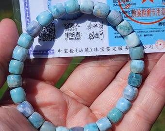 May include: A beaded bracelet featuring light blue and turquoise rectangular beads. The bracelet is held in a hand, with a certificate visible in the background. The beads have a slightly irregular shape and a polished finish.