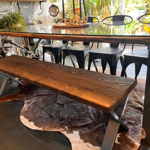May include: A rustic wooden bench with a dark brown stain and a metal frame. The bench is sitting on a cowhide rug in front of a glass-top table with black metal chairs.