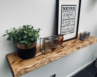 May include: A rustic wooden shelf with a burnt finish, featuring a potted succulent plant, decorative glass bottles, and two small glass candle holders. The shelf is mounted on a white wall with a framed print that reads "Never do anything by halves if you want to get away with it. Be outrageous. Go the whole hog. Make sure everything you do is completely crazy unbelievable."