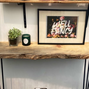 Könnte beinhalten: Ein rustikaler Holz-Konsolentisch mit schwarzen Haarnadelbeinen. Der Tisch hat ein Live-Edge-Design und ist mit einer grünen Kerze und einer kleinen Topfpflanze dekoriert. Ein gerahmter Druck mit dem Text "Well Fancy" ist auf dem Tisch ausgestellt.