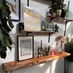 May include: A wooden shelf with a black metal frame, decorated with plants, a neon sign that reads "home sweet home", and a framed print with the text "Never do anything by halves if you want to get away with it. Be outrageous. Go the whole hog. Make sure everything you do is completely crazy unbelievable."
