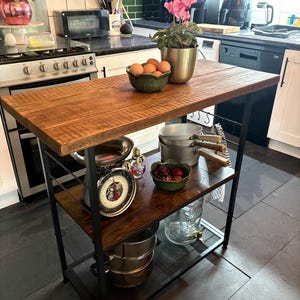 Puede incluir: Isla de cocina rústica con encimera de madera oscura y estructura de metal negro. La isla tiene dos estantes, con una balanza vintage, cuencos con huevos y fruta, y varios utensilios de cocina. Una planta en maceta se encuentra en la parte superior.