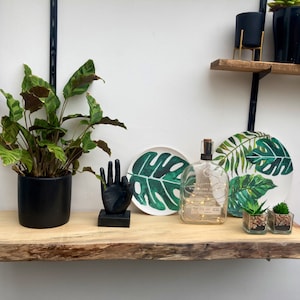 May include: A wooden shelf with a live plant, a black hand sculpture, two decorative plates with a tropical leaf design, a glass bottle with lights, and two small potted succulents.