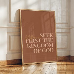 Può includere: Una stampa da parete rettangolare in una cornice di legno con sfondo color terracotta, che riporta il testo "SEEK FIRST THE KINGDOM OF GOD" in un carattere sans-serif chiaro e in grassetto, con la scritta in corsivo "Matthew 6:33" a sinistra.