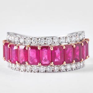 May include: Three stacked rings. The top and bottom rings are silver with small, clear stones. The middle ring is rose gold with rectangular, bright pink stones. The rings are set against a white background.