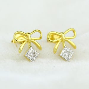 May include: A pair of gold stud earrings shaped like bows, each featuring a dangling charm set with a cluster of small, clear, round-cut stones. The metal has a polished yellow finish, and the stones are arranged in a circular pattern.