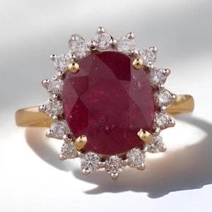 Op de afbeelding: Een gouden ring met een grote, ovaal geslepen robijn, omringd door een halo van kleine, ronde diamanten. De robijn heeft een dieprode kleur en de diamanten schitteren tegen het goud. Een klassiek en elegant sieraad.