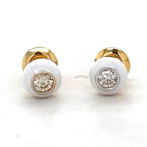 May include: A pair of stud earrings featuring a round white enamel setting with a central clear faceted stone. The base of each earring is crafted from polished yellow gold, providing a contrast to the white enamel and the sparkling center stone.