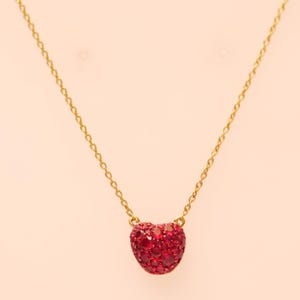May include: A gold-colored necklace featuring a heart-shaped pendant. The pendant is covered in small, vibrant red gemstones. The necklace chain is delicate and hangs against a soft, peach-colored background.