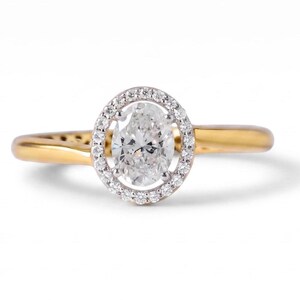May include: A gold ring featuring a central oval-cut clear gemstone surrounded by a halo of small round white stones. The band is smooth and polished gold, creating a continuous loop that supports the oval setting in the center of the piece.