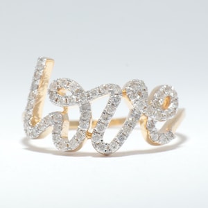 Op de afbeelding: Een gouden ring met het woord "love" in cursief geschreven. De letters zijn ingelegd met kleine, sprankelende diamanten. De ring staat tegen een witte achtergrond, wat het ontwerp benadrukt.