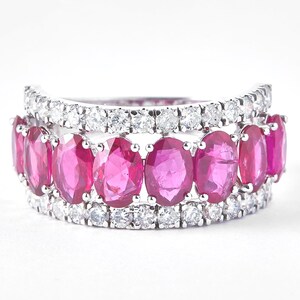 May include: A two-tiered ring featuring a row of oval-cut, vibrant pink gemstones set between rows of small, sparkling white gemstones. The ring has a bright, polished finish, and is set against a white background.