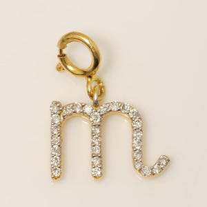 May include: A gold-colored charm in the shape of the letter "m", encrusted with small, clear gemstones. The charm has a clasp at the top for attachment to a necklace or bracelet. The gemstones sparkle against the gold.