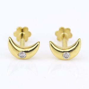 May include: A pair of gold stud earrings shaped like crescent moons. Each earring features a single small, clear, round-cut gemstone set into the center of the gold moon. The earrings have a post back with a flower-shaped backing mechanism.