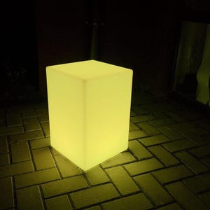 May include: A glowing, rectangular, yellow light cube stands on a brick surface. The cube emits a soft, warm light, creating a modern and ambient aesthetic. The cube is a solid, illuminated block.