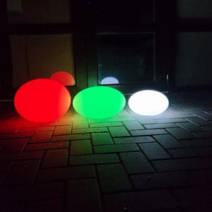 May include: Three glowing orbs in red, green, and white illuminate a dark outdoor setting. The orbs are different sizes and shapes, with the red one being a sphere and the green and white ones being oval. The orbs are placed on a brick surface.