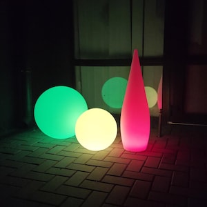 May include: A nighttime scene featuring illuminated outdoor decor. The image showcases a large green sphere, a smaller yellow sphere, and a tall, pink, teardrop-shaped light. Additional green and yellow spheres are visible in the background.