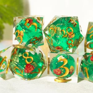 May include: A collection of clear resin dice with a green core, gold numerals, and decorative red strawberries and white flowers. The dice are in various shapes, including a cube, a d20, and a sphere.