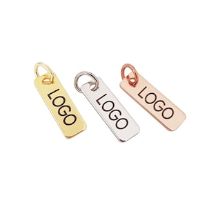 Könnte beinhalten: Drei Metallanhänger mit einem Ring oben. Die Anhänger sind rechteckig und haben das Wort "LOGO" eingraviert. Die Anhänger sind gold, silber und roségold.